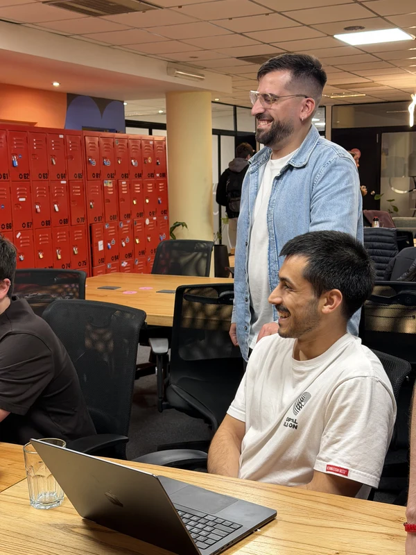 A teammate standing to talk with a seated colleague working on a laptop