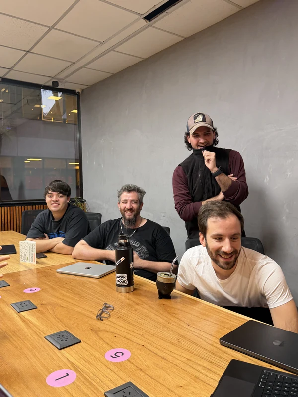 Five colleagues laughing around a meeting table covered in laptops and drinks
